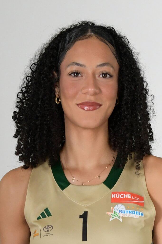 Eine Frau mit lockigen Haaren lächelt im sandfarbenen Kader Damen 1 Basketballtrikot mit der Nummer 1 und mehreren Sponsorenlogos